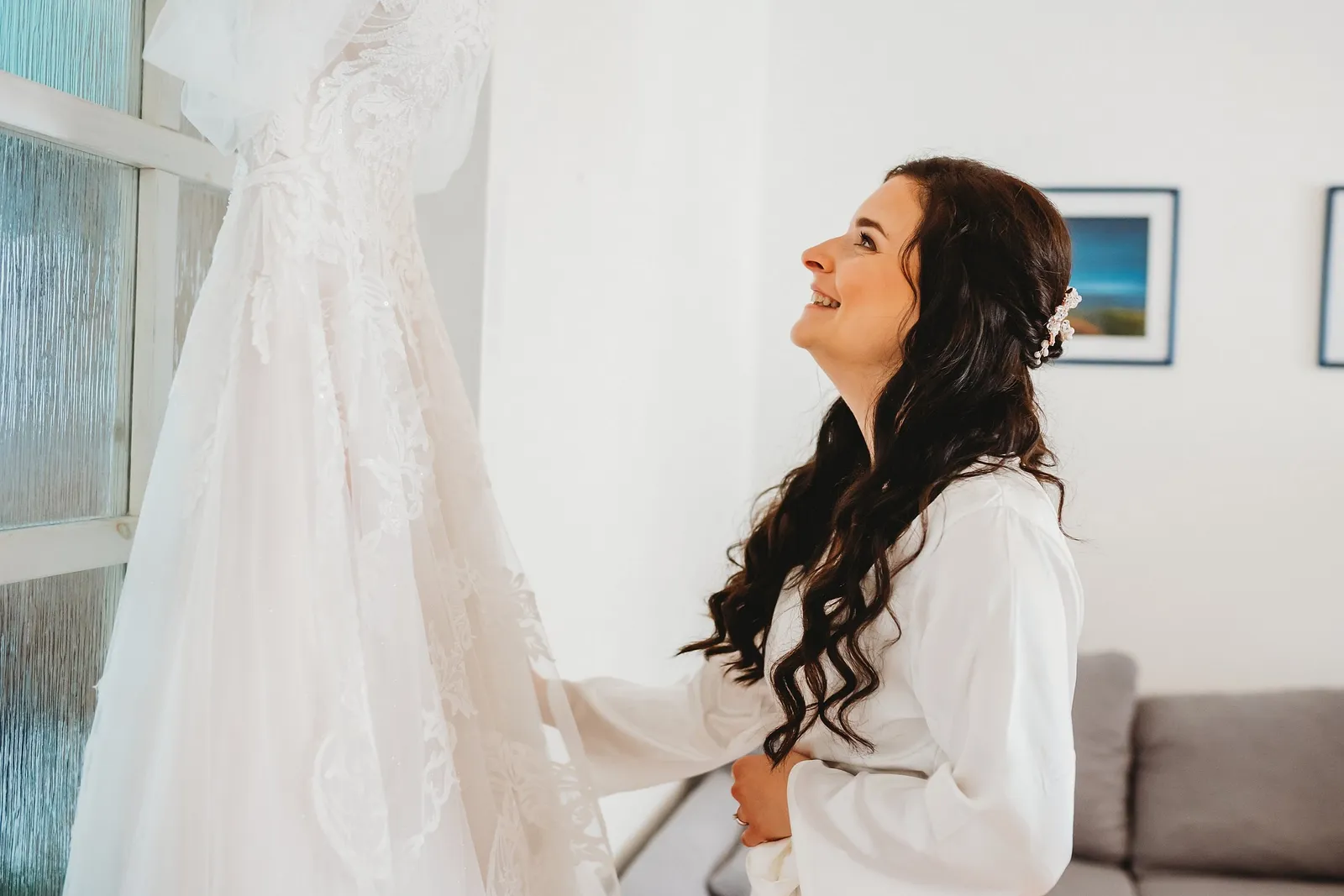 Usmiata nevesta v bielom župane s ozdobou vo vlasoch hľadí na čipkované svadobné šaty zavesené na okne – svadba Majka a Marek, Hotel Máj, Liptovský Ján