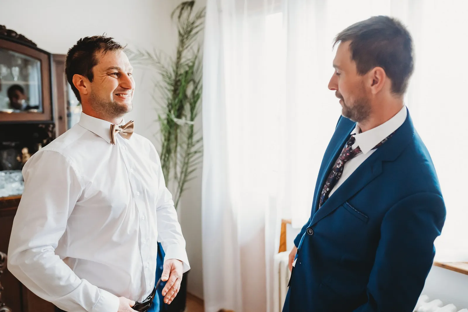 Rozosmiaty ženích s béžovým motýlikom sa pozerá na muža v modrom obleku pri okne so záclonou – svadba Majka a Marek, Hotel Máj, Liptovský Ján