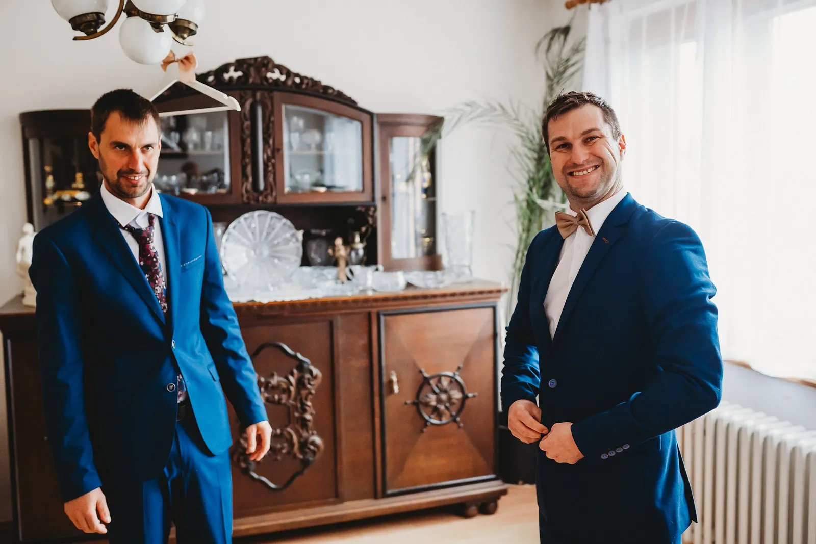 Ženích si zapína modré sako, vedľa neho stojí muž v obleku s kvetovanou kravatou pri vyrezávanom kredenci – svadba Majka a Marek, Hotel Máj, Liptovský Ján