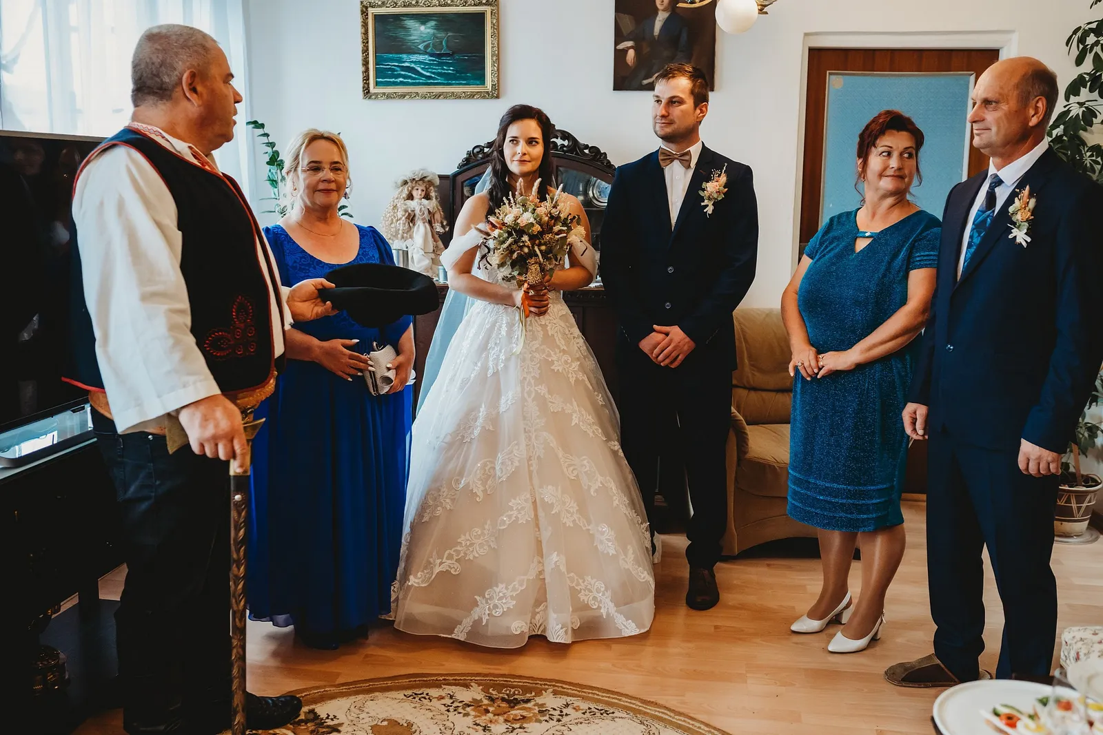 Nevesta s kyticou a ženích s motýlikom počúvajú starejšieho s klobúkom v obývačke medzi rodičmi – svadba Majka a Marek, Hotel Máj, Liptovský Ján