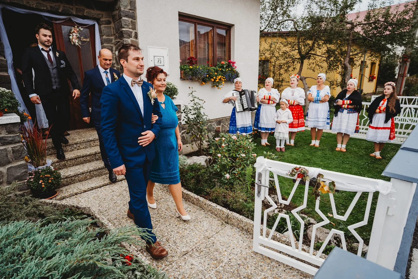 Ženích vychádza z domu pod ruku so ženou v modrom, na trávniku spievajú ženy v krojoch s harmonikou – svadba Majka a Marek, Hotel Máj, Liptovský Ján