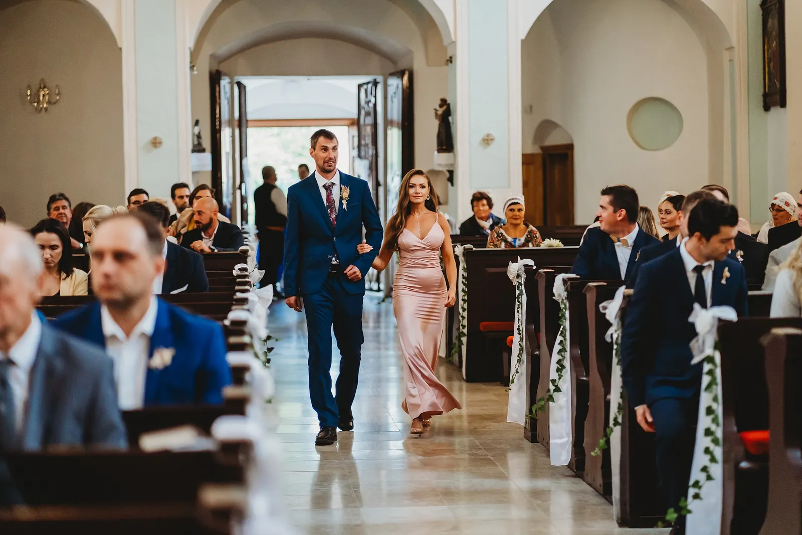 Svedok v modrom obleku kráča uličkou kostola s družičkou v ružových saténových šatách – svadba Majka a Marek, Hotel Máj, Liptovský Ján