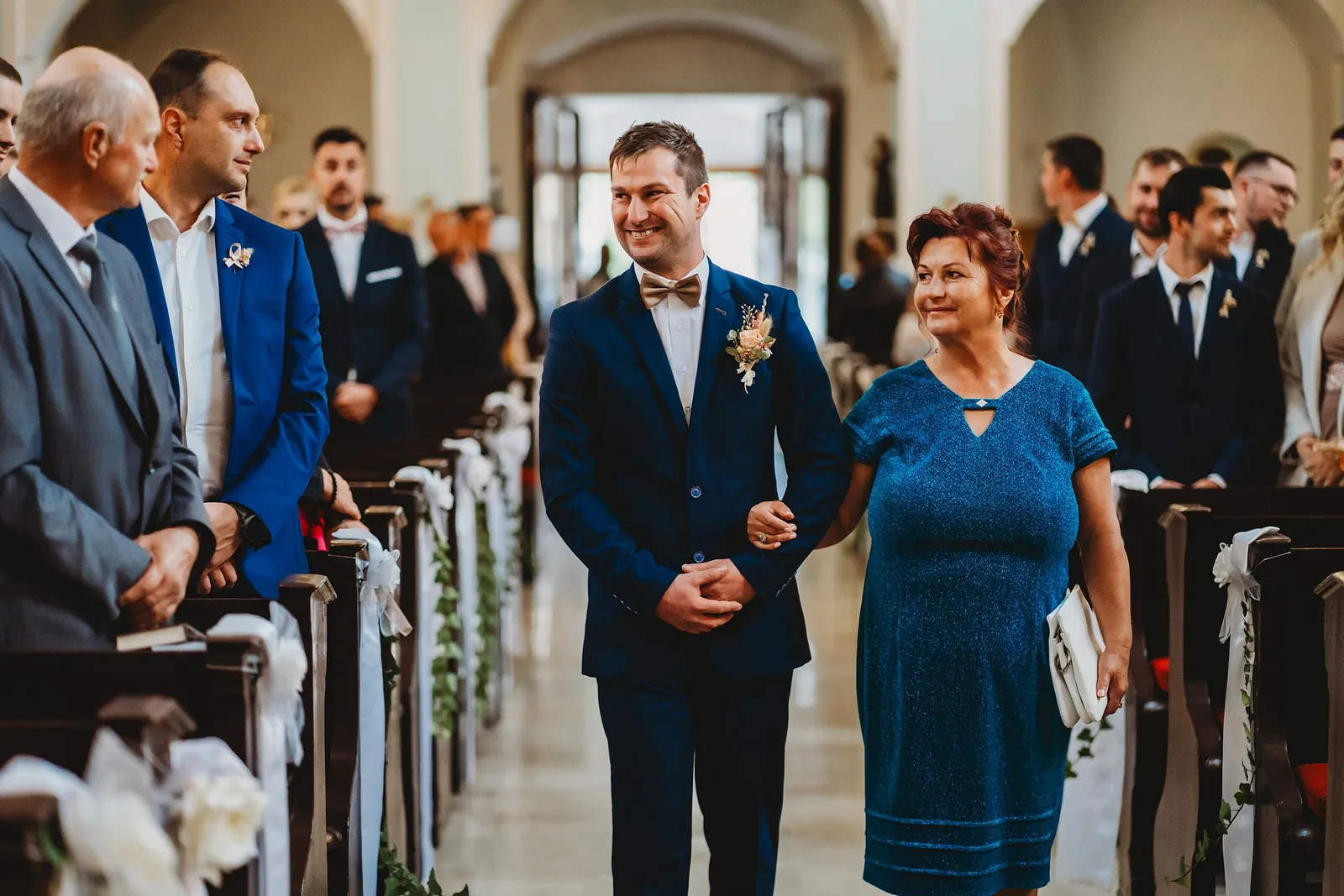 Usmiaty ženích s motýlikom vedie uličkou kostola ženu v modrých šatách popri laviciach s hosťami – svadba Majka a Marek, Hotel Máj, Liptovský Ján