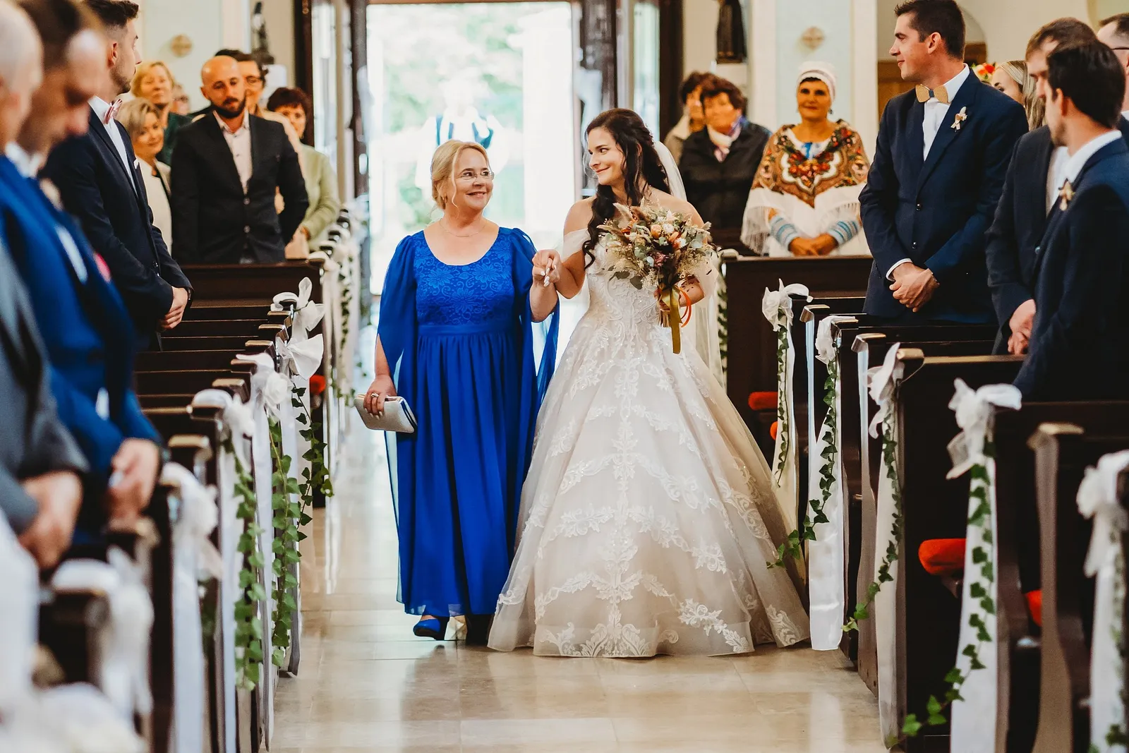 Nevesta s jesennou kyticou kráča k oltáru za ruku so ženou v modrých šatách, hostia stoja v laviciach – svadba Majka a Marek, Hotel Máj, Liptovský Ján