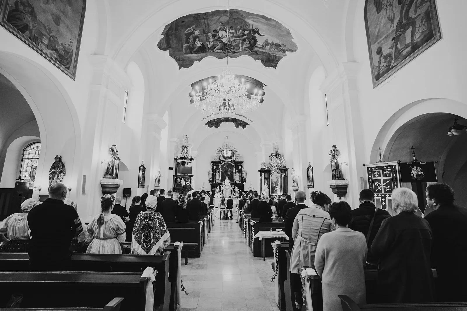 Čiernobiely pohľad od vchodu do barokového kostola s freskami, lustrom a stojacimi hosťami – svadba Majka a Marek, Hotel Máj, Liptovský Ján