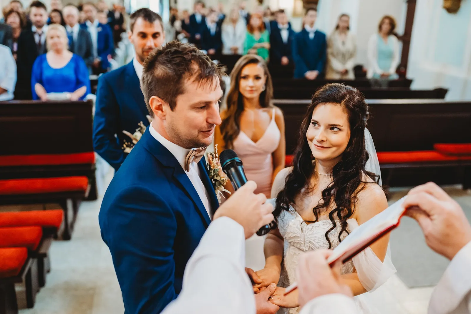 Ženích hovorí manželský sľub do mikrofónu, ktorý drží kňaz, nevesta sa naňho nežne pozerá – svadba Majka a Marek, Hotel Máj, Liptovský Ján