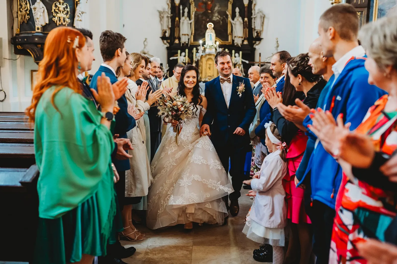 Novomanželia vychádzajú uličkou kostola, hostia im po oboch stranách tlieskajú, vpredu malé dievčatko – svadba Majka a Marek, Hotel Máj, Liptovský Ján