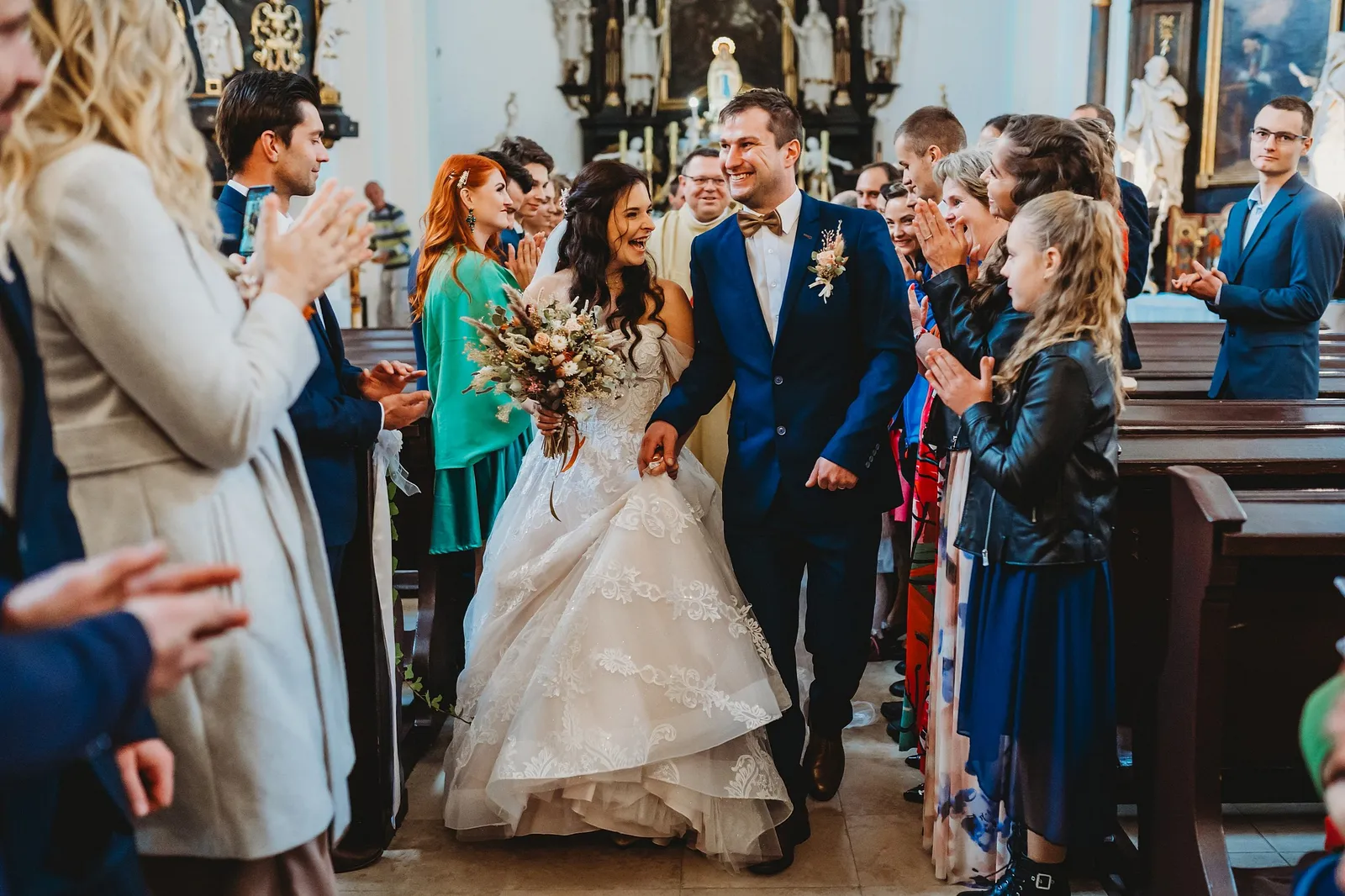 Rozosmiata nevesta a ženích sa držia za ruky v uličke kostola, dievčatko v koženej bunde tlieska – svadba Majka a Marek, Hotel Máj, Liptovský Ján