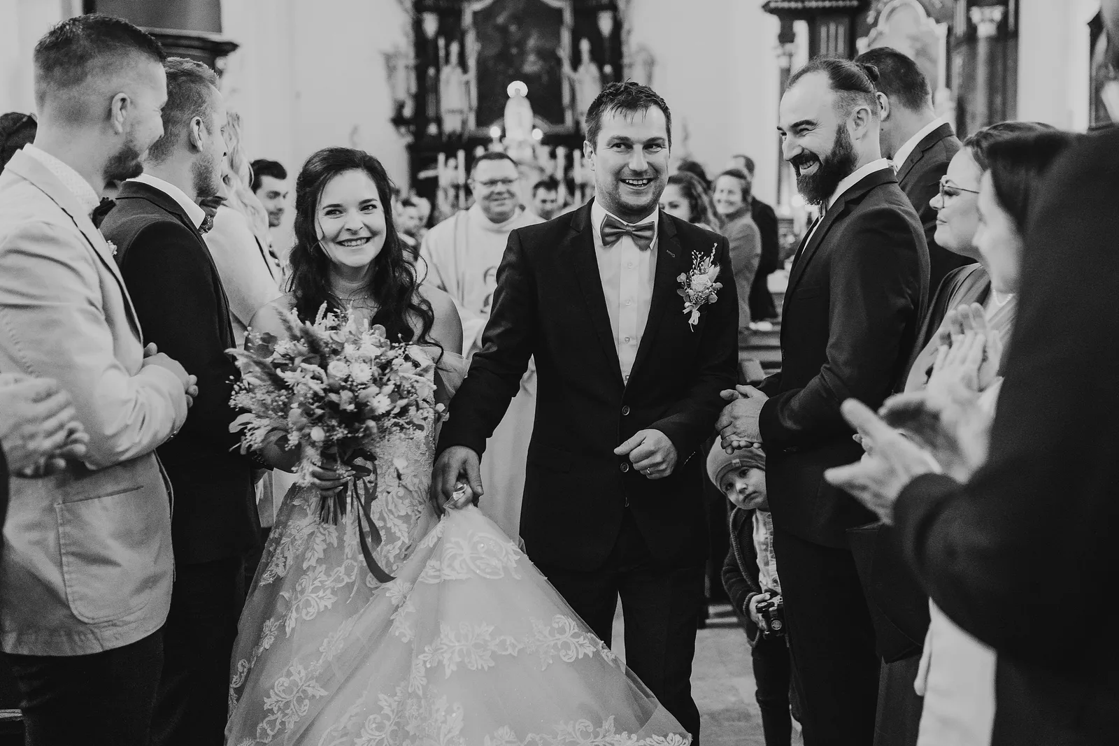 Čiernobiely odchod novomanželov z kostola, bradatý hosť sa na nich smeje a ostatní tlieskajú – svadba Majka a Marek, Hotel Máj, Liptovský Ján