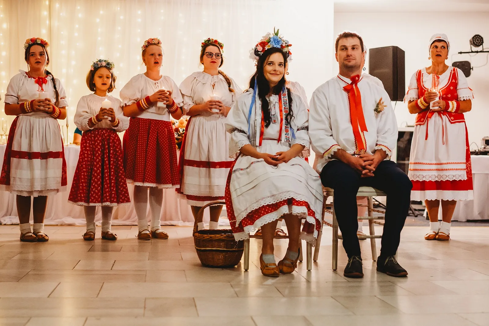 Novomanželia v krojoch sedia pri čepčení, za nimi ženy a dievčatá v krojoch so sviečkami – svadba Majka a Marek, Hotel Máj, Liptovský Ján