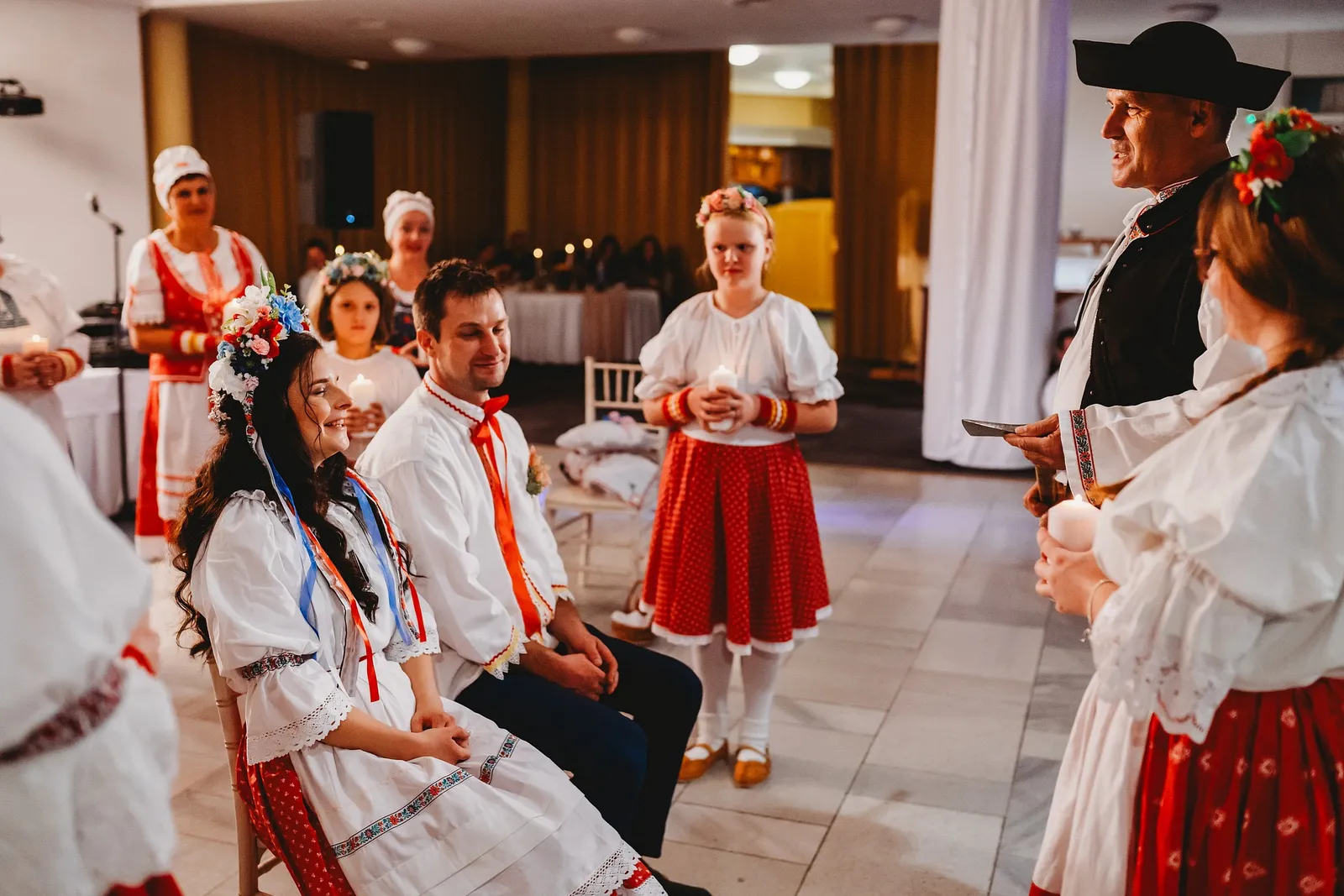 Usmiata nevesta s vencom z kvetov a ženích v kroji sedia, kým starejší číta z mobilu – svadba Majka a Marek, Hotel Máj, Liptovský Ján