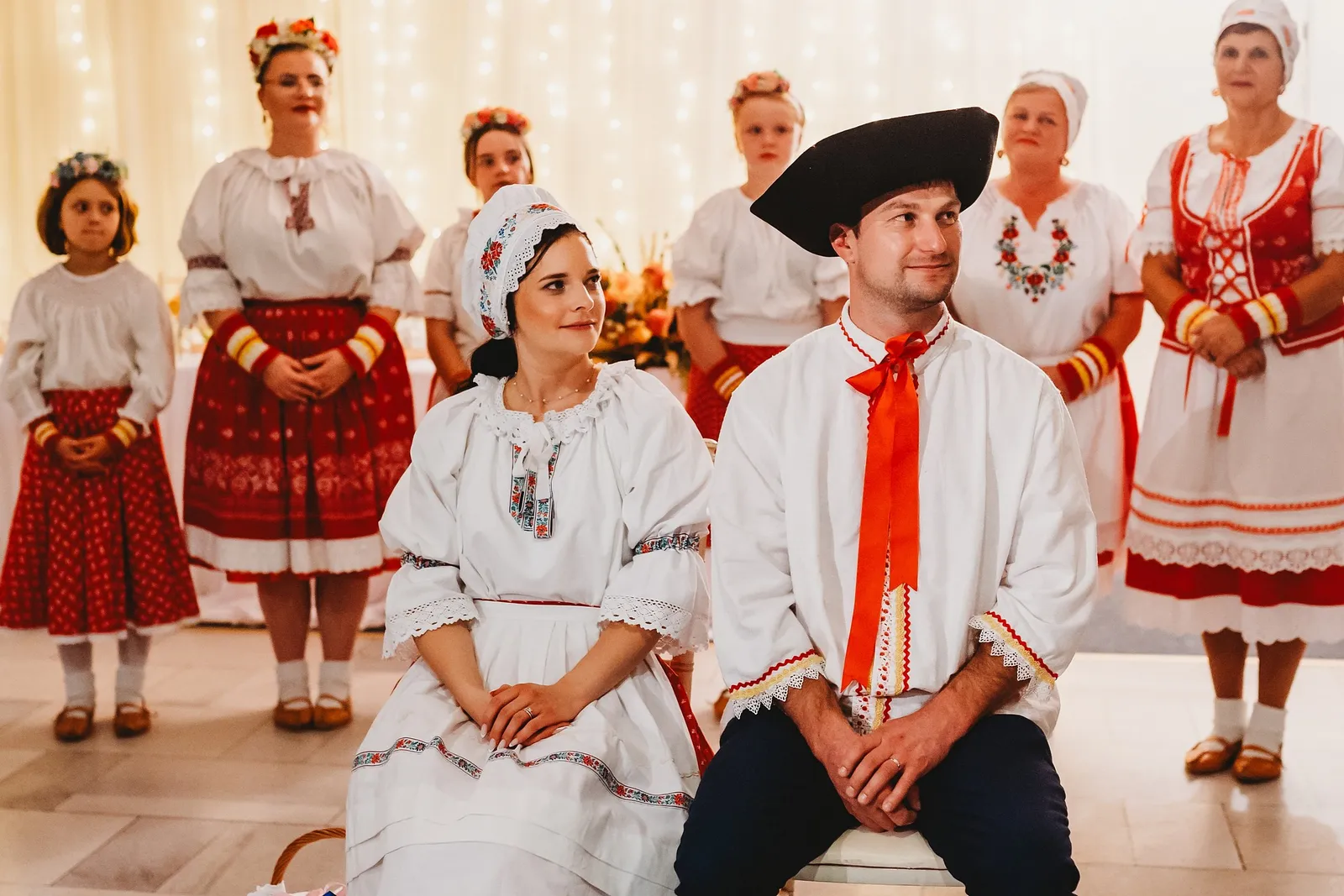 Nevesta v čepci a ženích v klobúku s červenou stuhou sedia pred ženami v krojoch – svadba Majka a Marek, Hotel Máj, Liptovský Ján