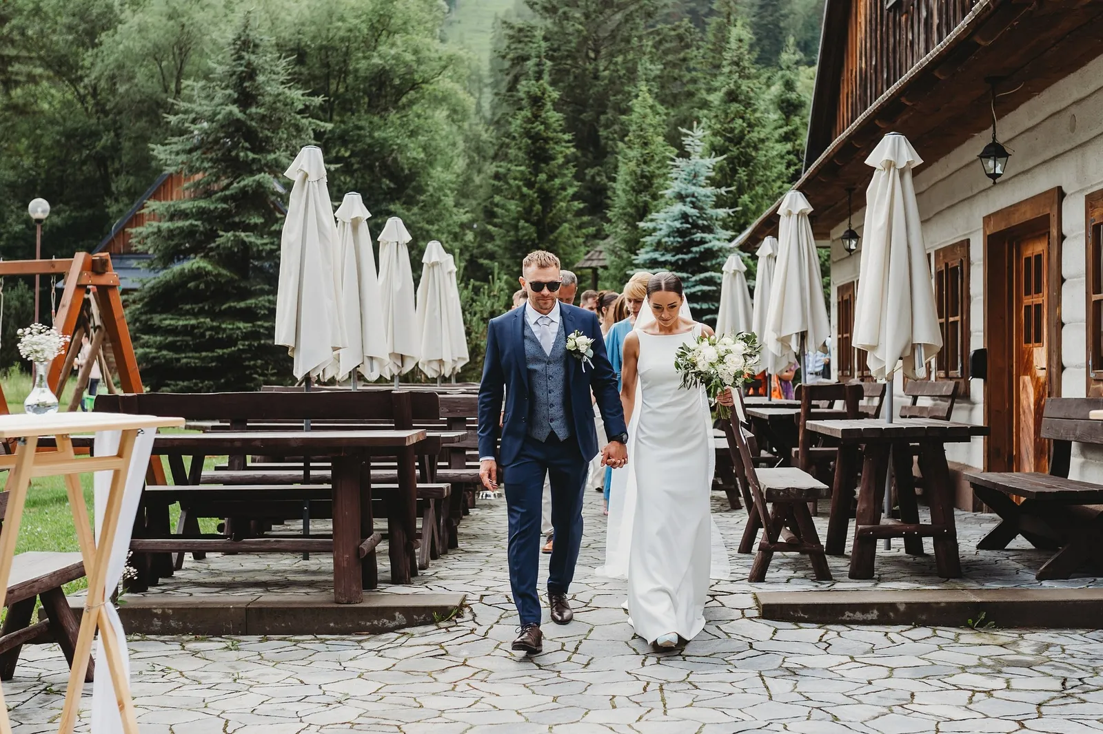 Novomanželia ruka v ruke prichádzajú na kamennú terasu so zloženými slnečníkmi a drevenicami – svadba Pajka a Majo, Stodola Liptovský dvor