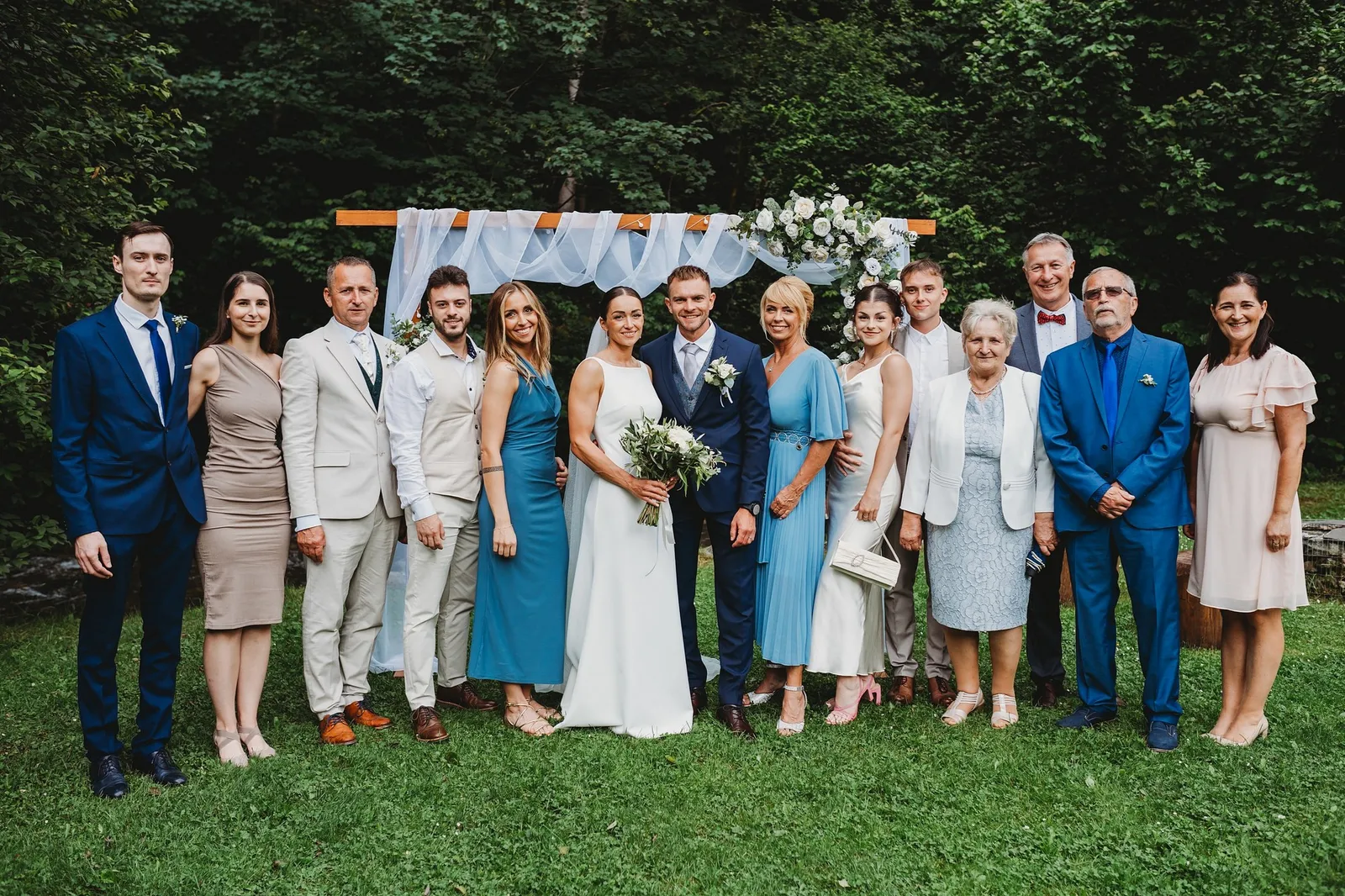 Spoločná fotografia novomanželov s rodinou na tráve pred drevenou slávobránou s bielou látkou – svadba Pajka a Majo, Stodola Liptovský dvor