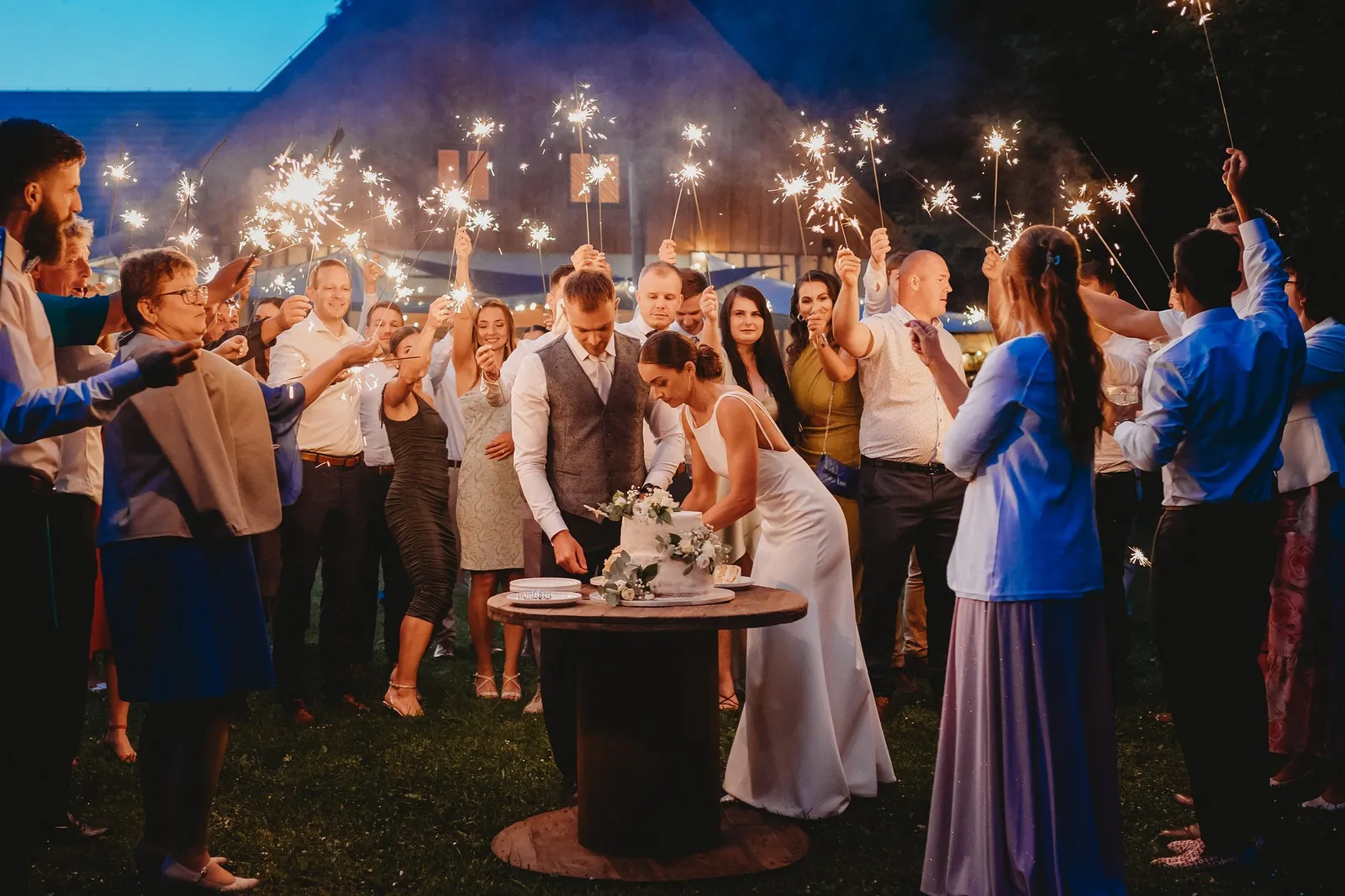 Nevesta a ženích pri krájaní torty za súmraku, obklopení svadobčanmi so zapálenými prskavkami – svadba Pajka a Majo, Stodola Liptovský dvor