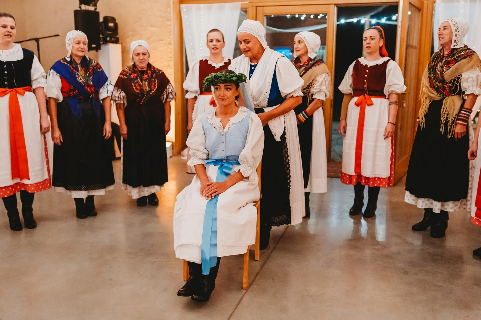 Čepčenie – nevesta v kroji s venčekom zo zelene sedí na stoličke medzi ženami vo folklórnych krojoch – svadba Pajka a Majo, Stodola Liptovský dvor