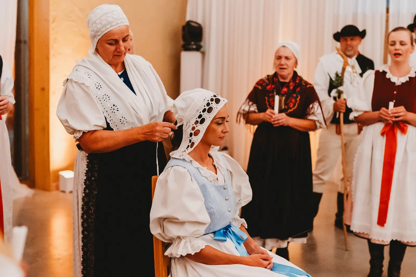 Žena v kroji nasadzuje neveste čipkovaný čepiec, za nimi stoja speváčky so sviečkami – svadba Pajka a Majo, Stodola Liptovský dvor