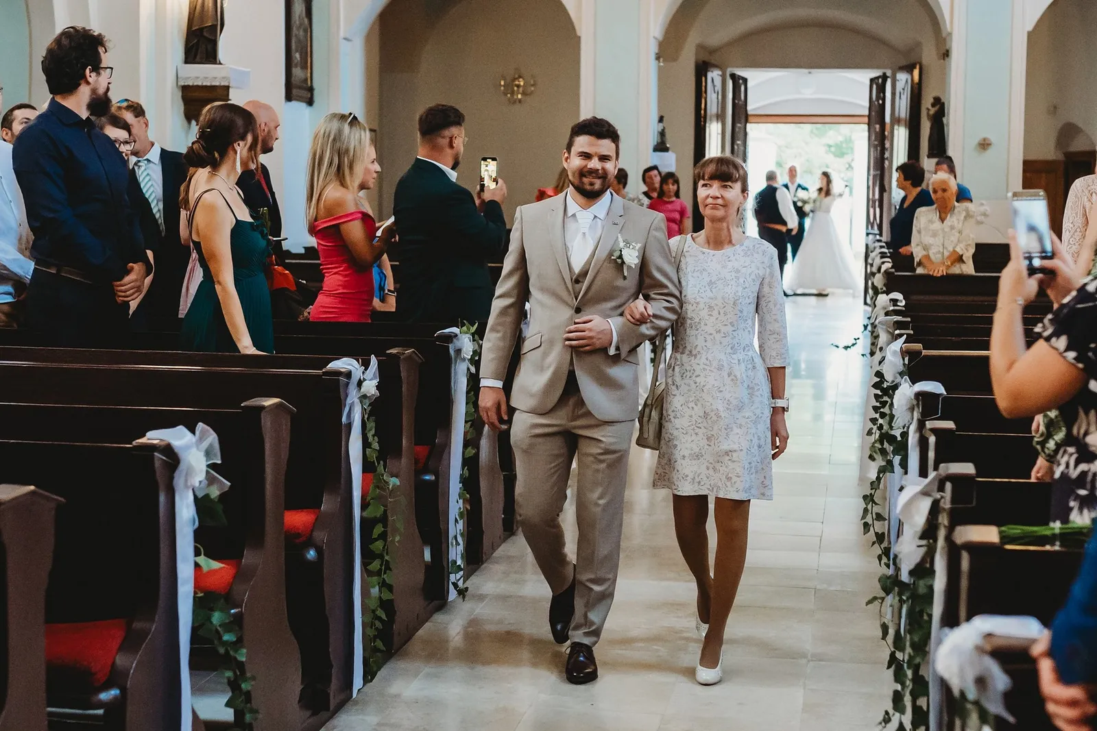 Ženích kráča uličkou kostola so ženou v čipkových šatách, v pozadí vchádza nevesta – svadba Peťka a Peťo, Stodola Liptovský dvor