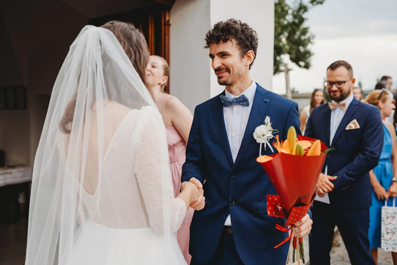 Nevesta podáva ruku hosťovi v modrom obleku s motýlikom, ktorý drží oranžovú kyticu – svadba Peťka a Peťo, Stodola Liptovský dvor