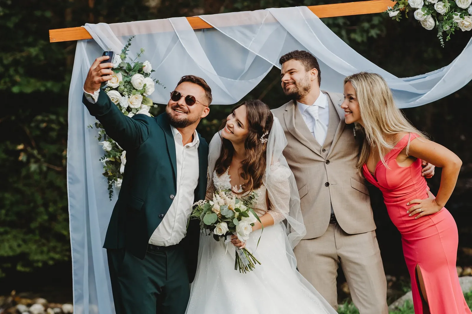 Hosť v slnečných okuliaroch si robí selfie s novomanželmi a hosťkou v ružových šatách pri kvetinovej bráne – svadba Peťka a Peťo, Stodola Liptovský dvor