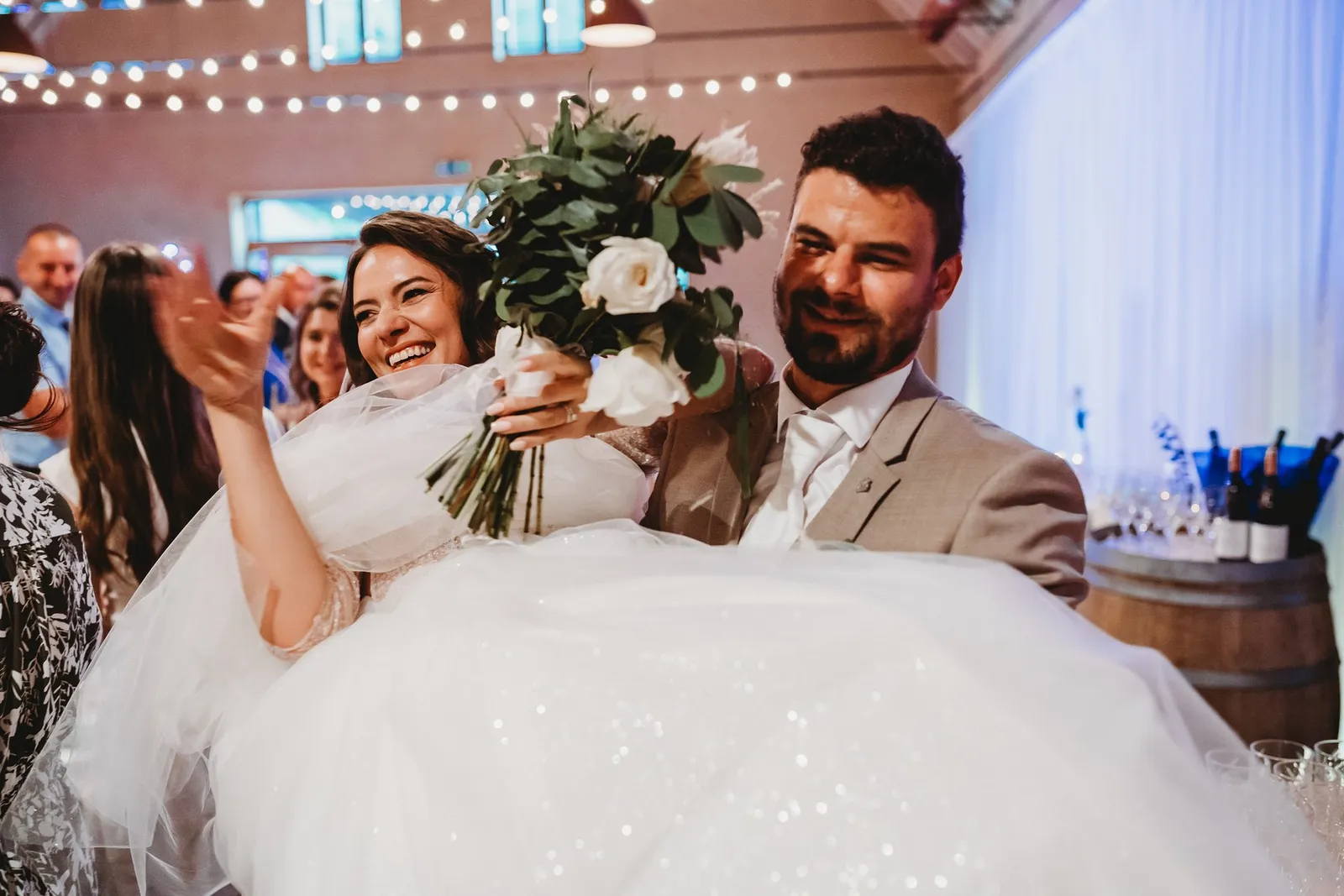 Ženích nesie rozosmiatu nevestu s kyticou na rukách do sály plnej svetielok – svadba Peťka a Peťo, Stodola Liptovský dvor