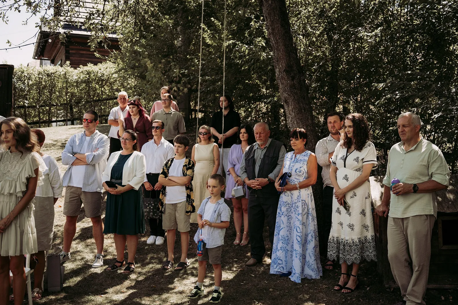 Hostia rôzneho veku stoja v tieni stromov a pozorne sledujú svadobný obrad – svadba Ivka a Ondro, Humno Trnka, Záskov