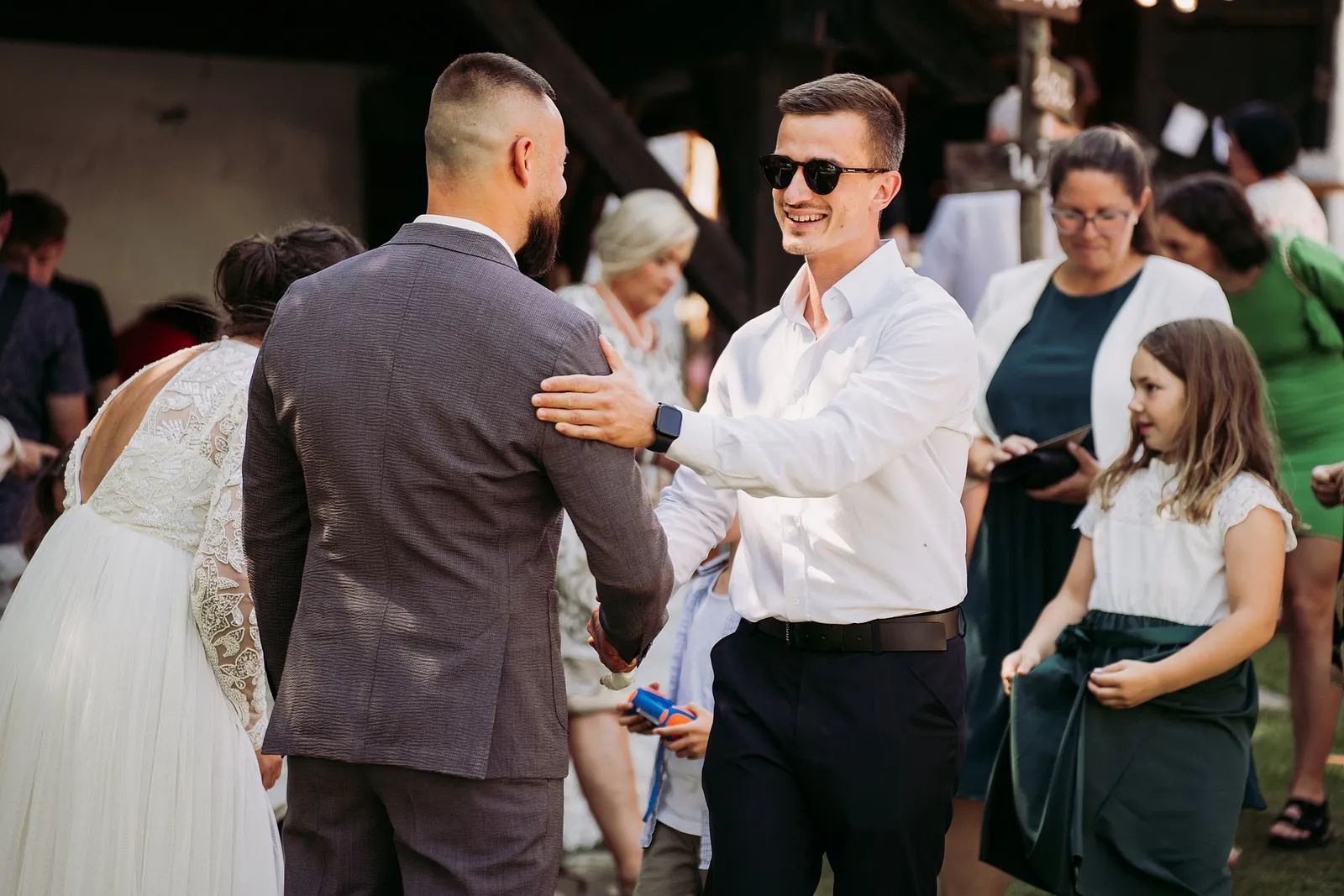 Hosť v slnečných okuliaroch s úsmevom gratuluje ženíchovi a poklepáva ho po ramene – svadba Ivka a Ondro, Humno Trnka, Záskov