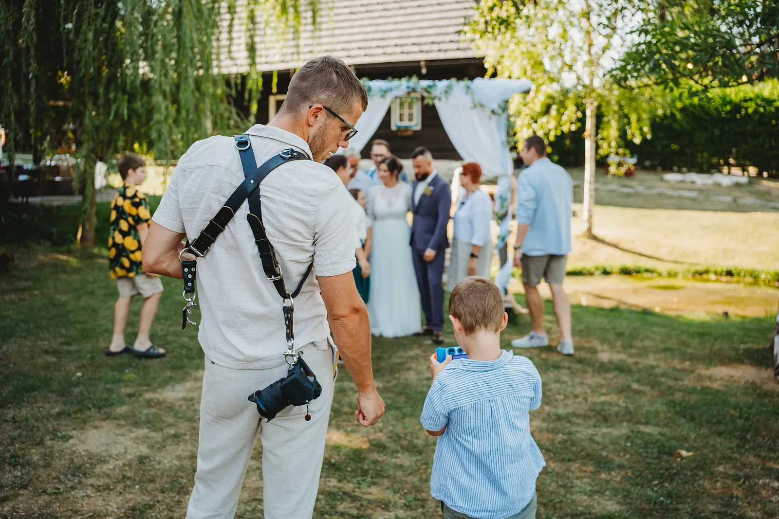 Malý chlapec s foťákom napodobňuje fotografa pri obrade na záhrade – svadba Ivka a Ondro, Humno Trnka, Záskov