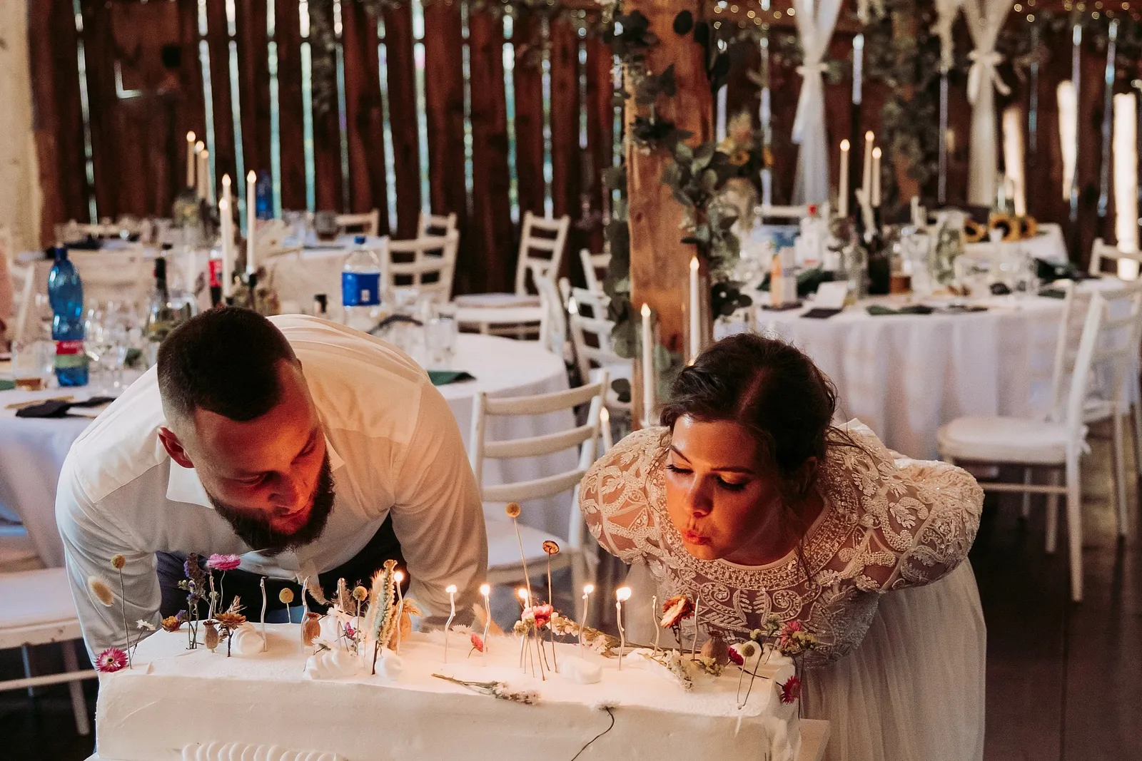 Nevesta a ženích sfukujú sviečky na nízkej bielej torte s kvetmi – svadba Ivka a Ondro, Humno Trnka, Záskov