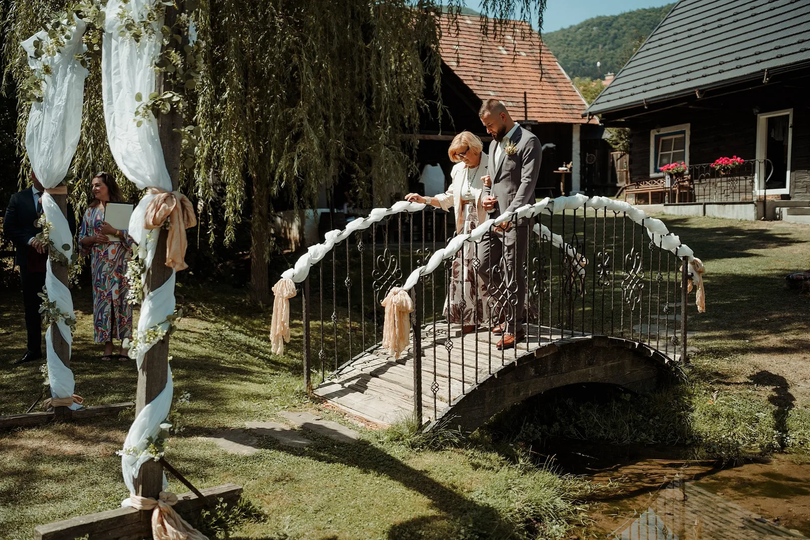 Ženích vedie staršiu dámu cez ozdobený kovový mostík k obradnej bráne pod vŕbou – svadba Ivka a Ondro, Humno Trnka, Záskov