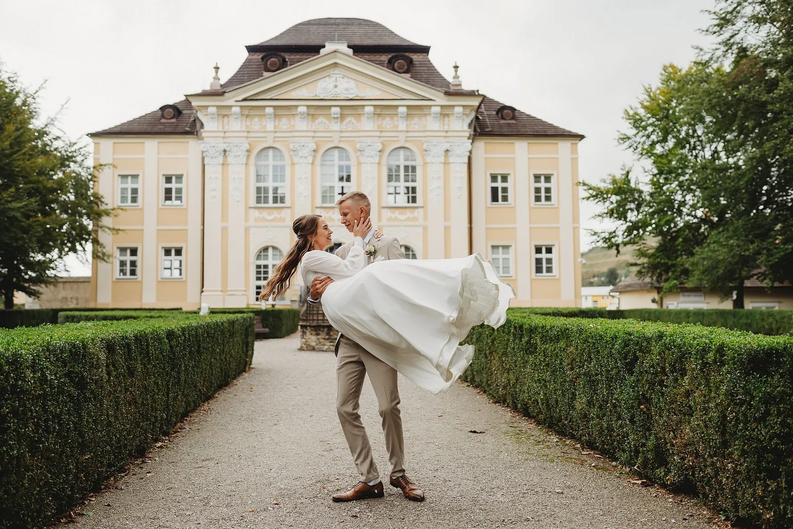 Ženích roztáča nevestu na rukách v aleji so strihanými živými plotmi pred barokovým kaštieľom