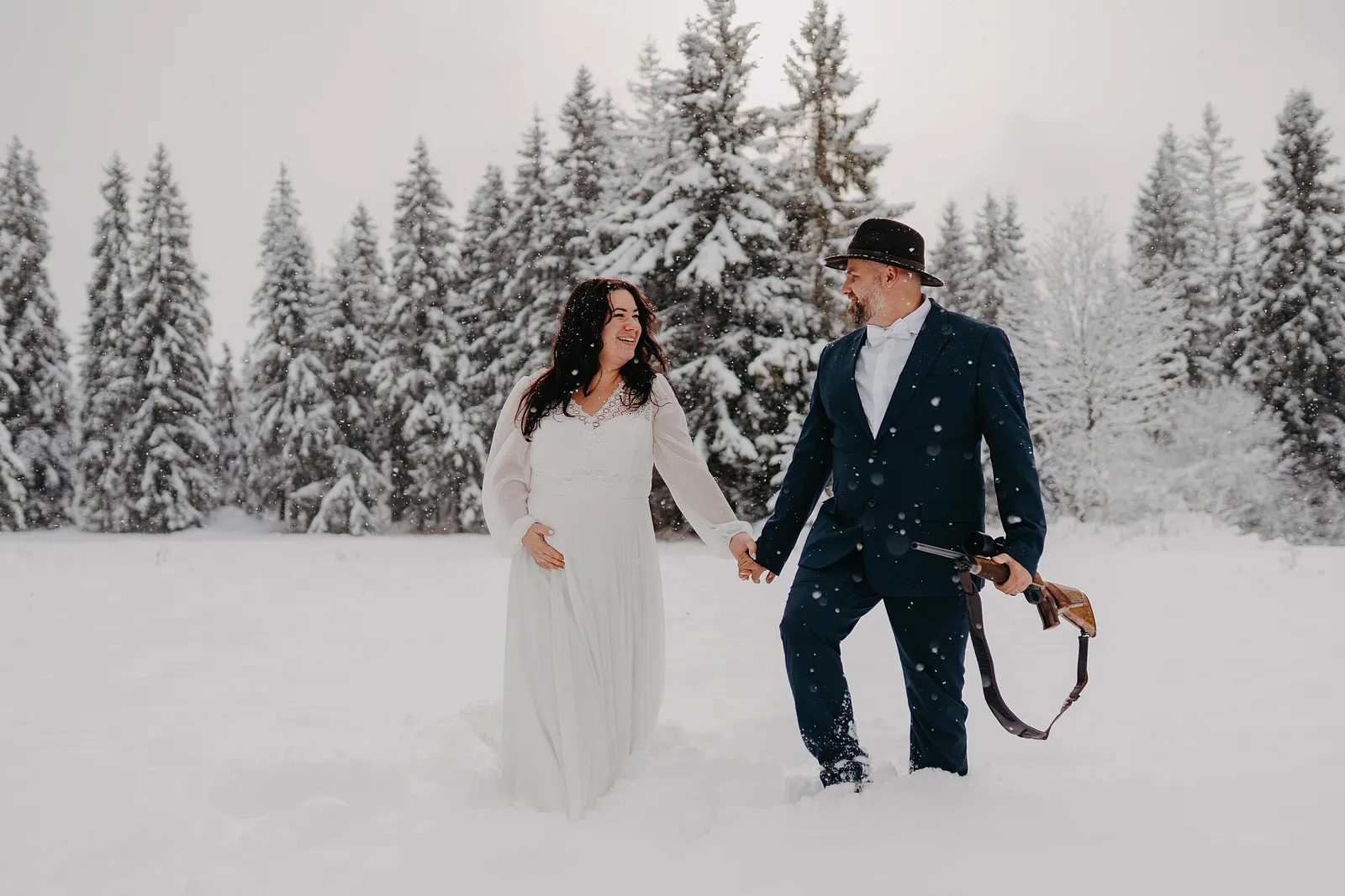 Zimná svadobná fotografia nevesty a ženícha v klobúku, ktorí sa držia za ruky v snehu medzi zasneženými smrekmi