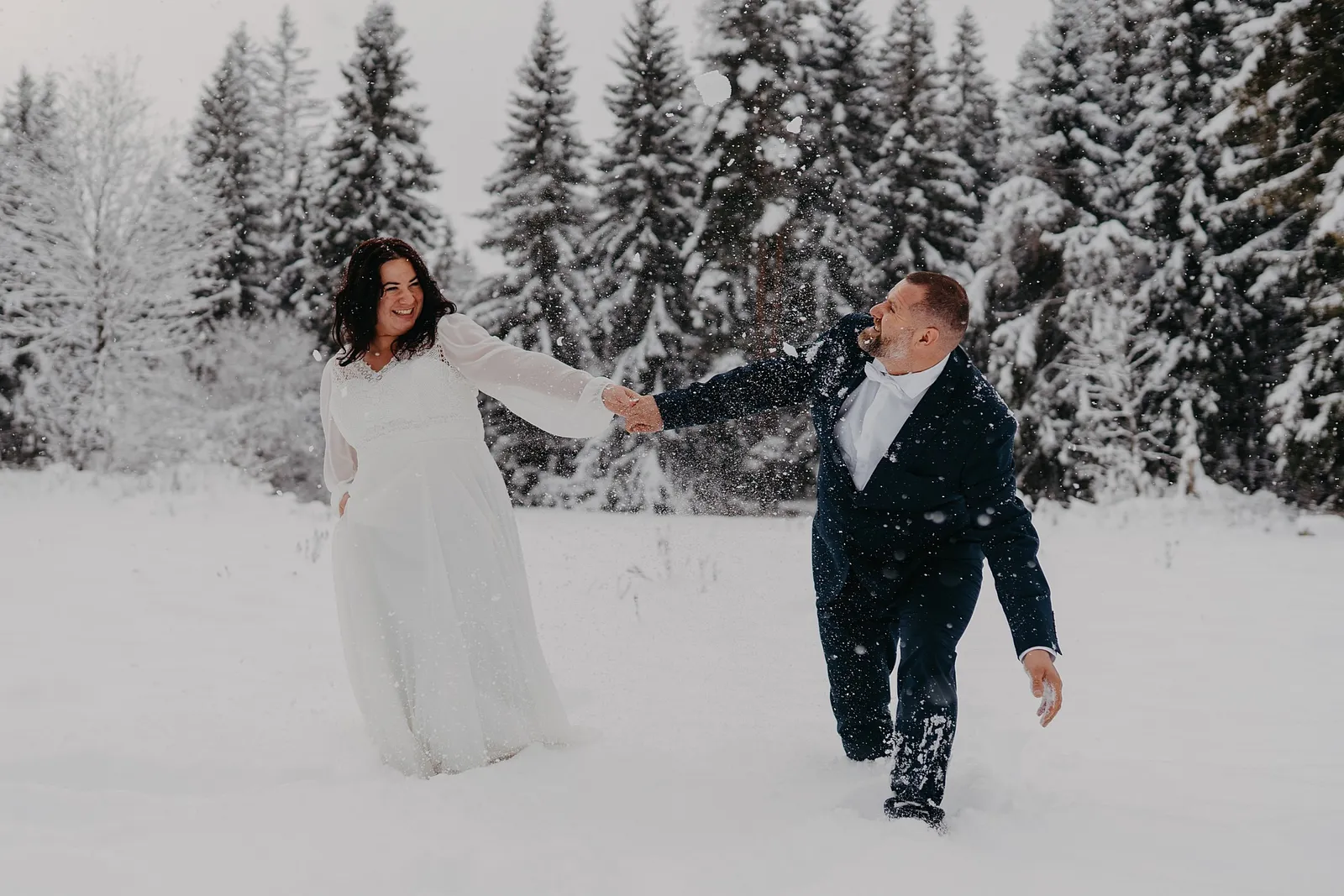 Rozosmiata nevesta ťahá ženícha za ruku hlbokým snehom pred zasneženým smrekovým lesom