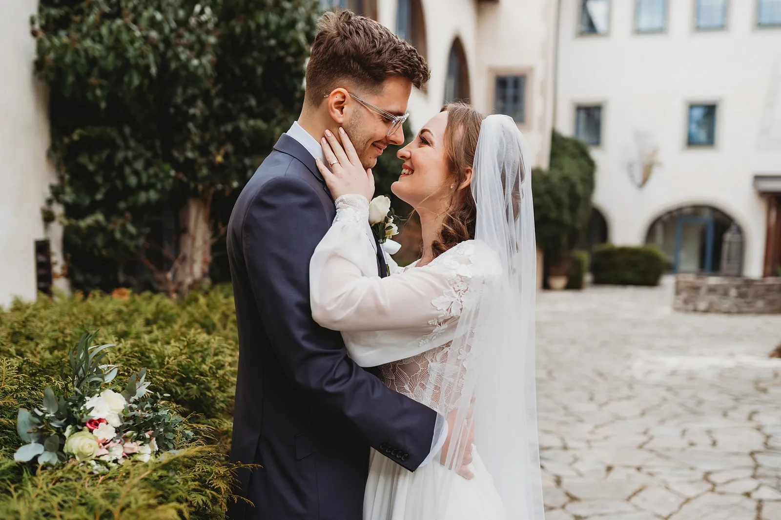 Nevesta so závojom hladí po tvári ženícha v okuliaroch, objímajú sa na nádvorí, v tráve leží biela kytica