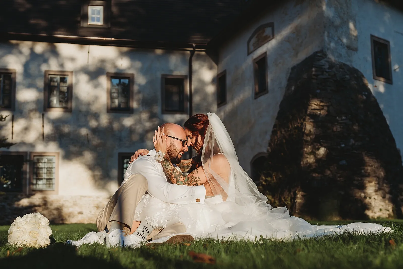 Nevesta so závojom a tetovaný ženích sedia objatí na tráve pred starou budovou, vedľa nich biela kytica