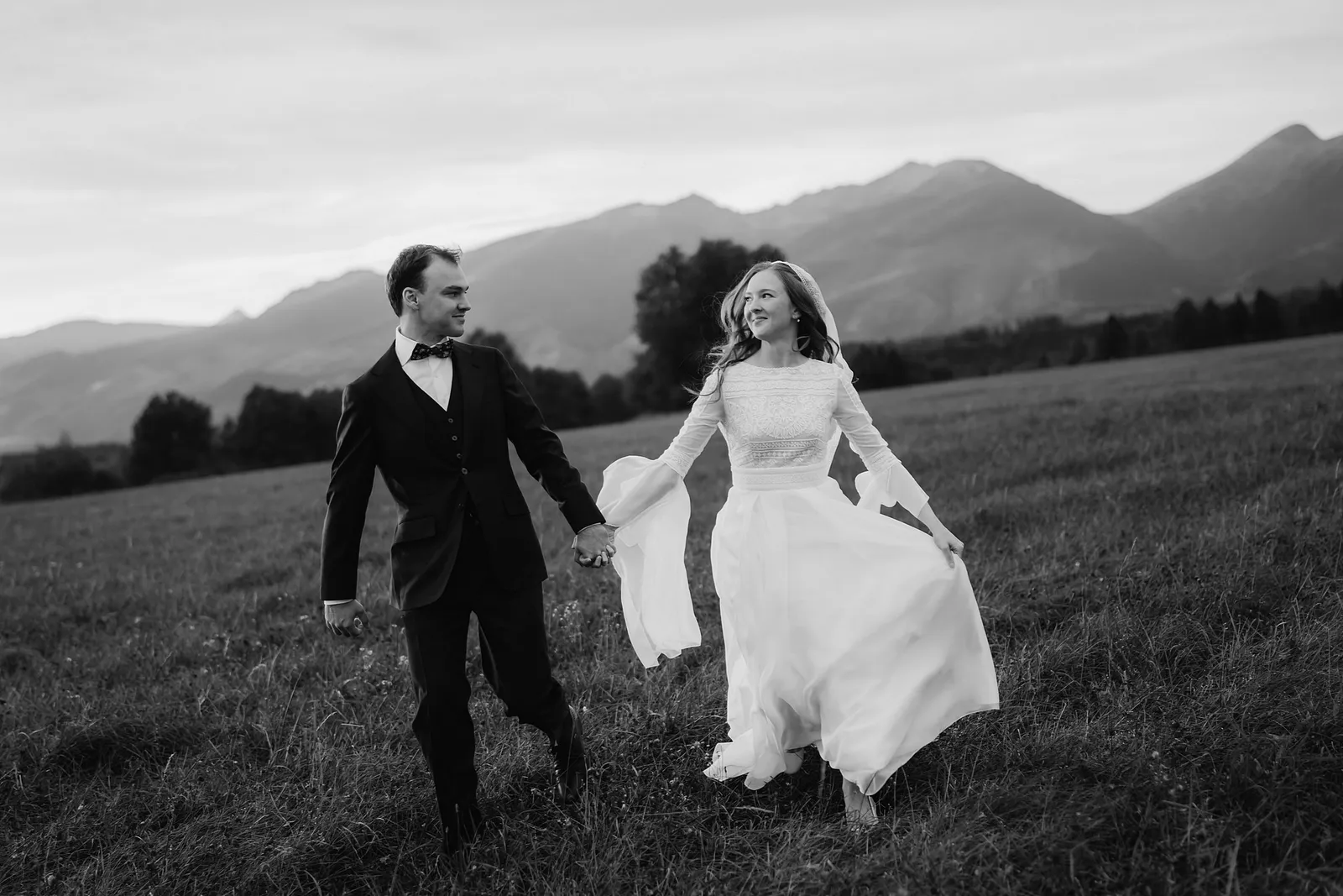 Čiernobiela svadobná fotografia páru, ktorý beží ruka v ruke po lúke, za nimi pohorie v oparu
