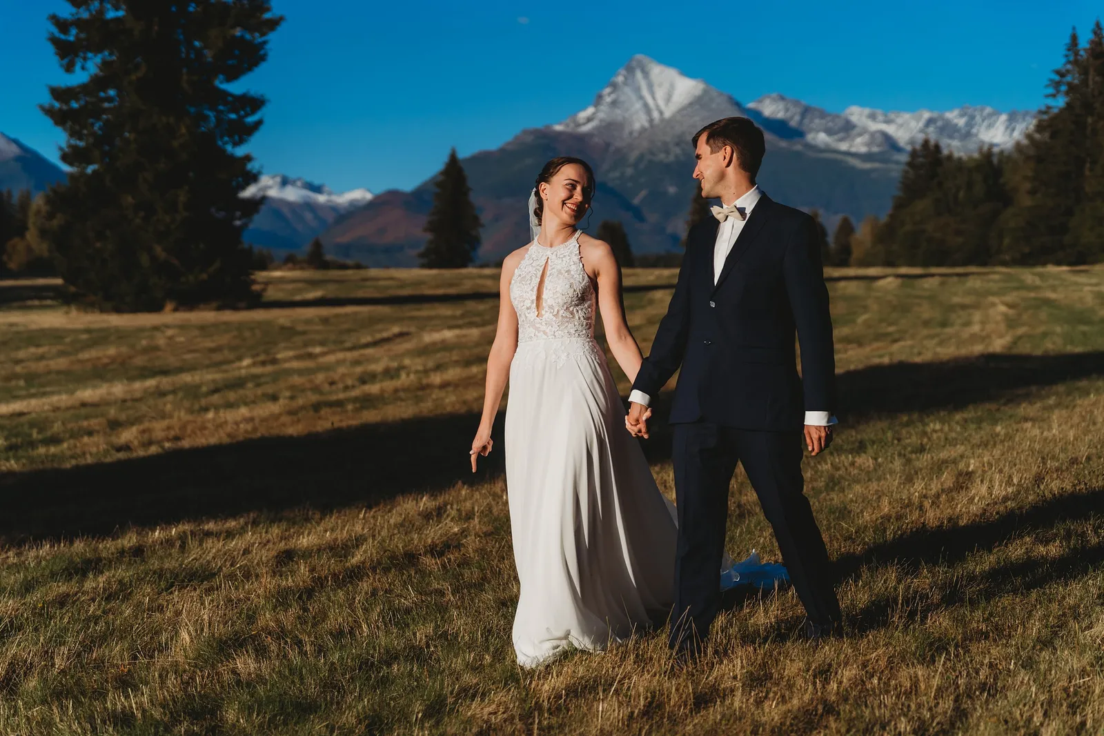Usmiata nevesta vedie ženícha za ruku po jesennej lúke, v pozadí zasnežené štíty hôr