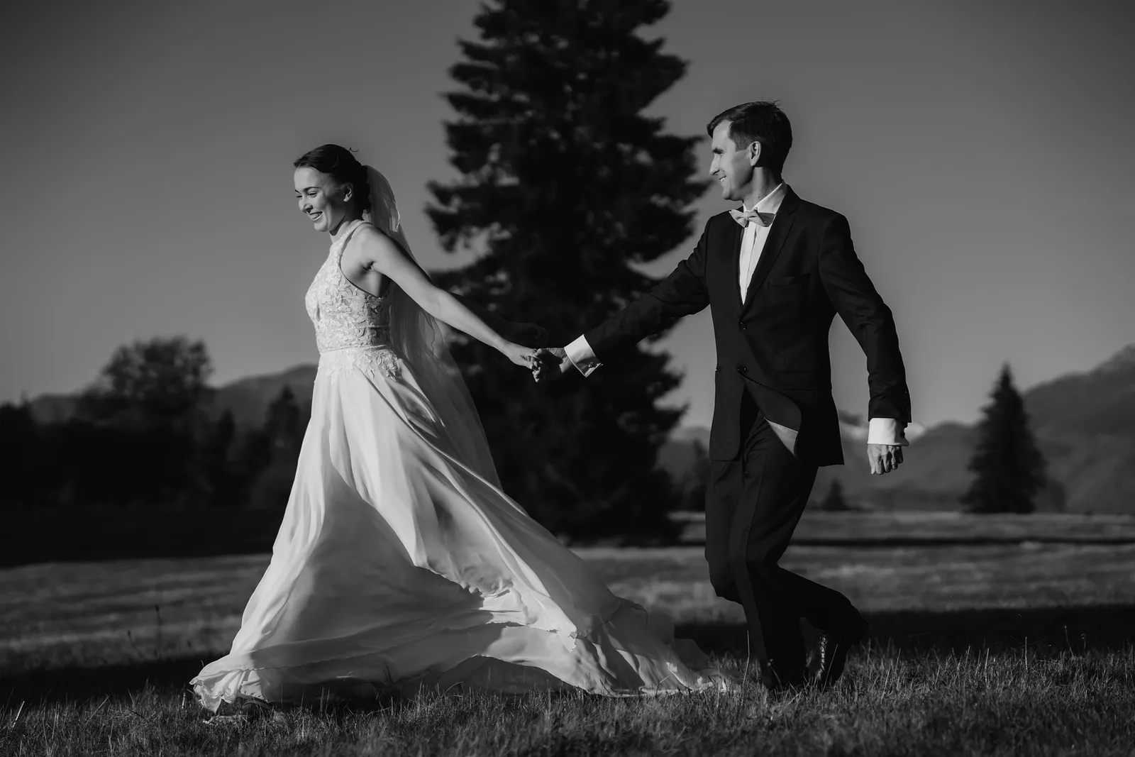 Čiernobiela svadobná fotografia novomanželov, ktorí sa držia za ruky a bežia po lúke pri smreku