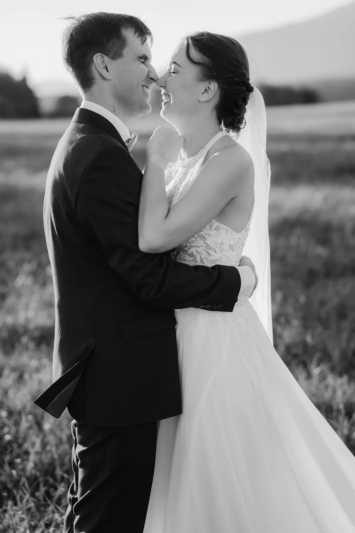Čiernobiela fotografia nevesty a ženícha, ktorí sa s úsmevom dotýkajú nosmi v objatí na lúke