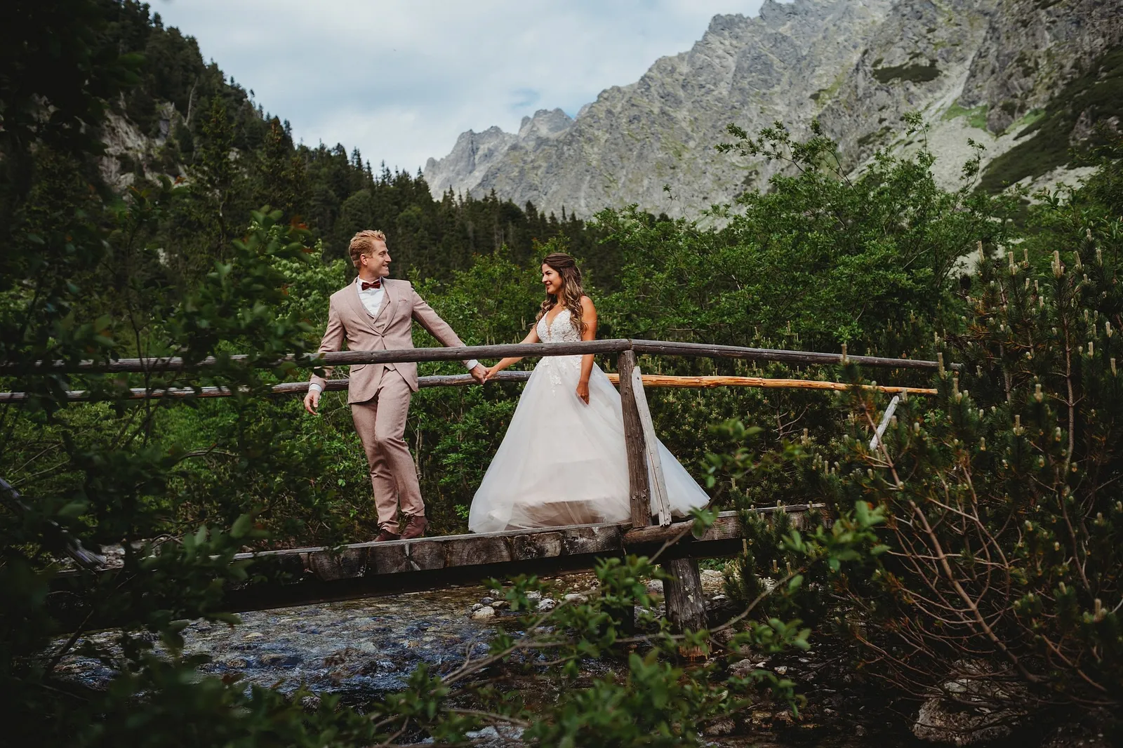 Ženích v béžovom obleku vedie nevestu cez drevený mostík nad horským potokom, za nimi skalnaté štíty