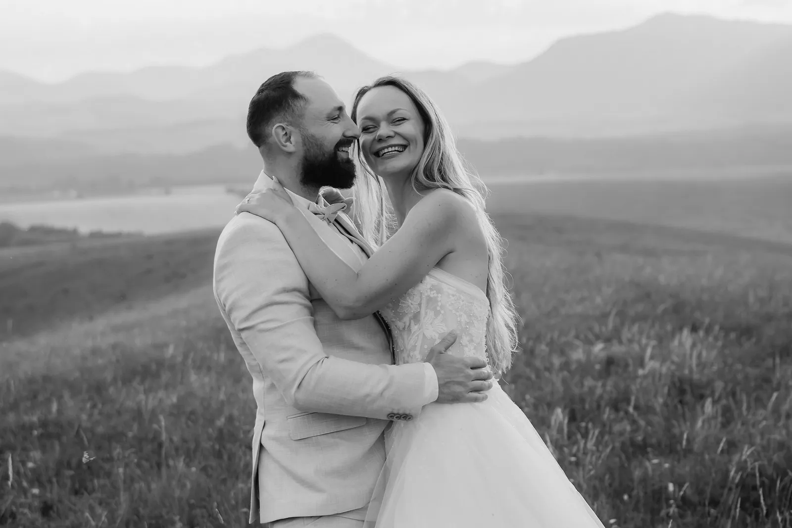 Čiernobiela svadobná fotografia smejúcej sa nevesty, ktorá sa túli k ženíchovi na kopci nad jazerom