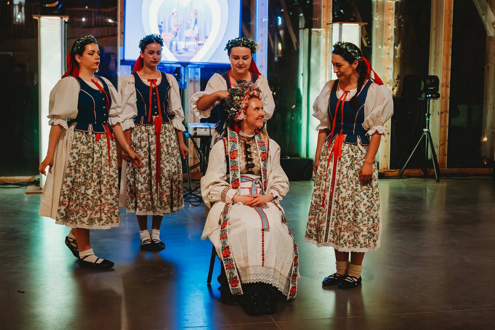 Čepčenie nevesty, ktorá sedí v kroji obklopená ženami v ľudových krojoch s vencami – svadba Sandra a Sebastian, Stodola Gréta