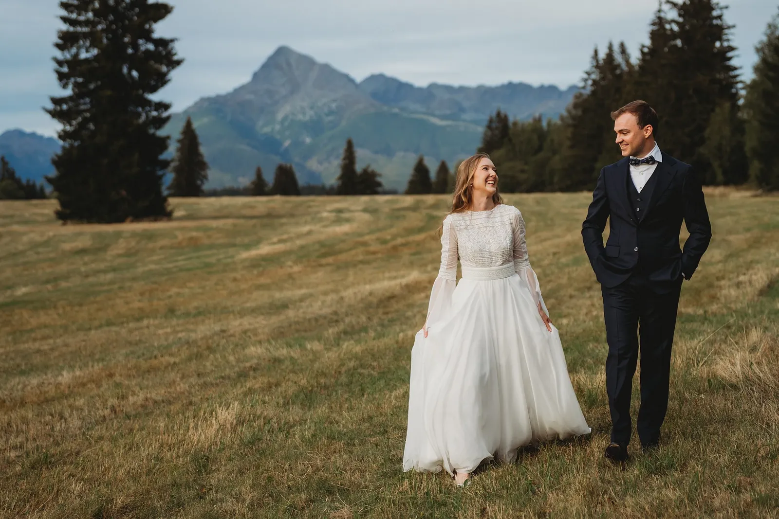 Nevesta a ženích sa prechádzajú lúkou a usmievajú sa na seba, za nimi smreky a horský štít – svadba Sandra a Sebastian, Stodola Gréta