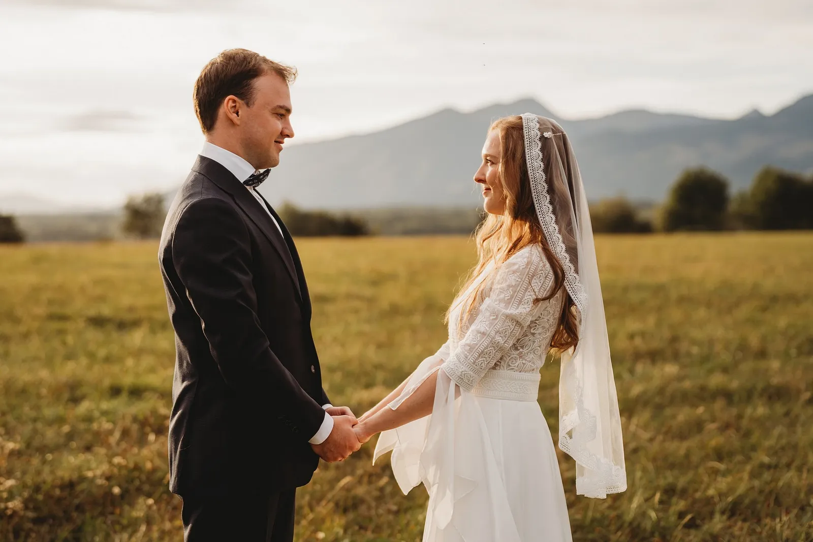 Pár sa drží za ruky tvárou v tvár na lúke v zlatom svetle, nevesta s dlhým čipkovým závojom – svadba Sandra a Sebastian, Stodola Gréta