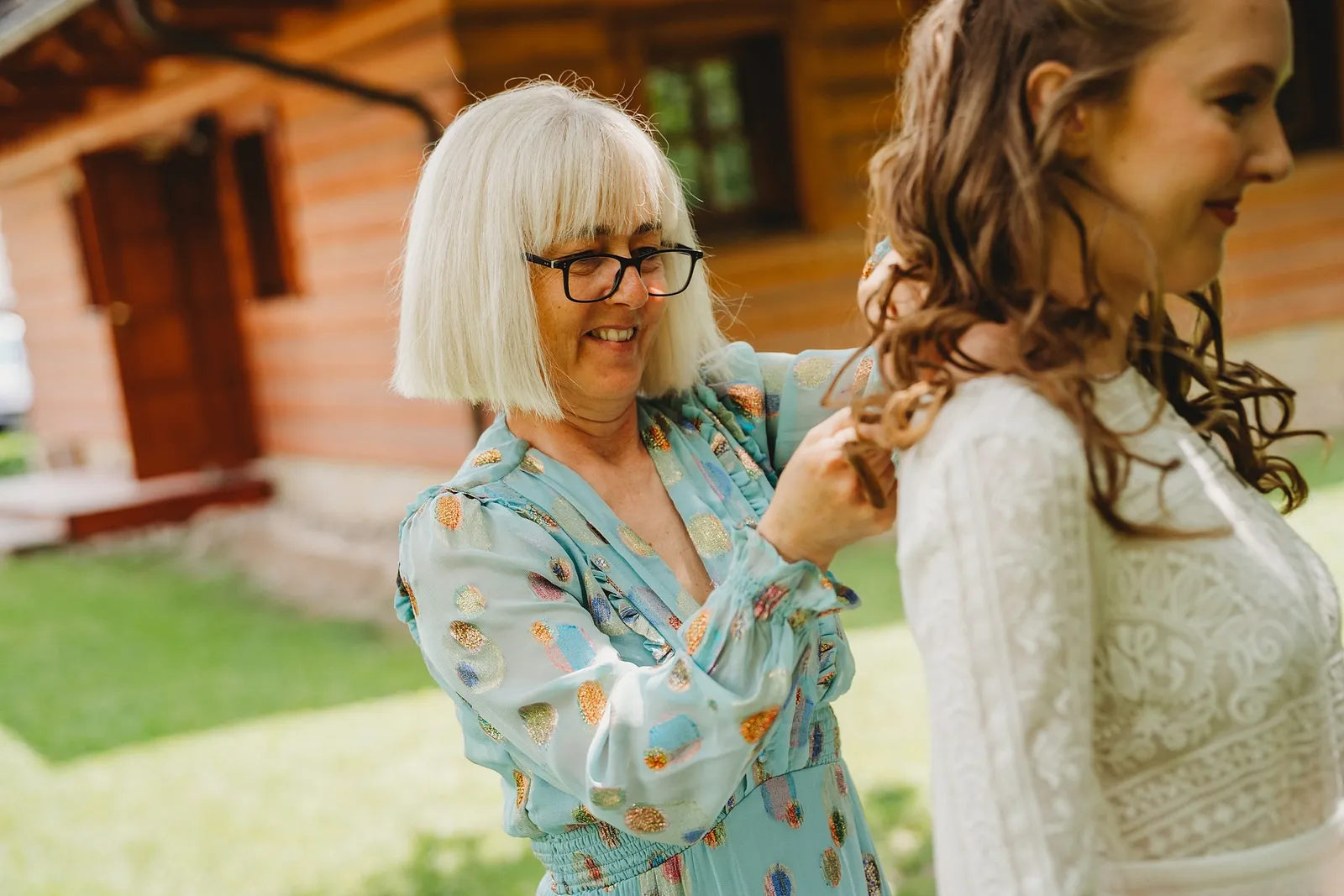 Žena s bielymi vlasmi a okuliarmi upravuje neveste šaty pred dreveným zrubom – svadba Sandra a Sebastian, Stodola Gréta