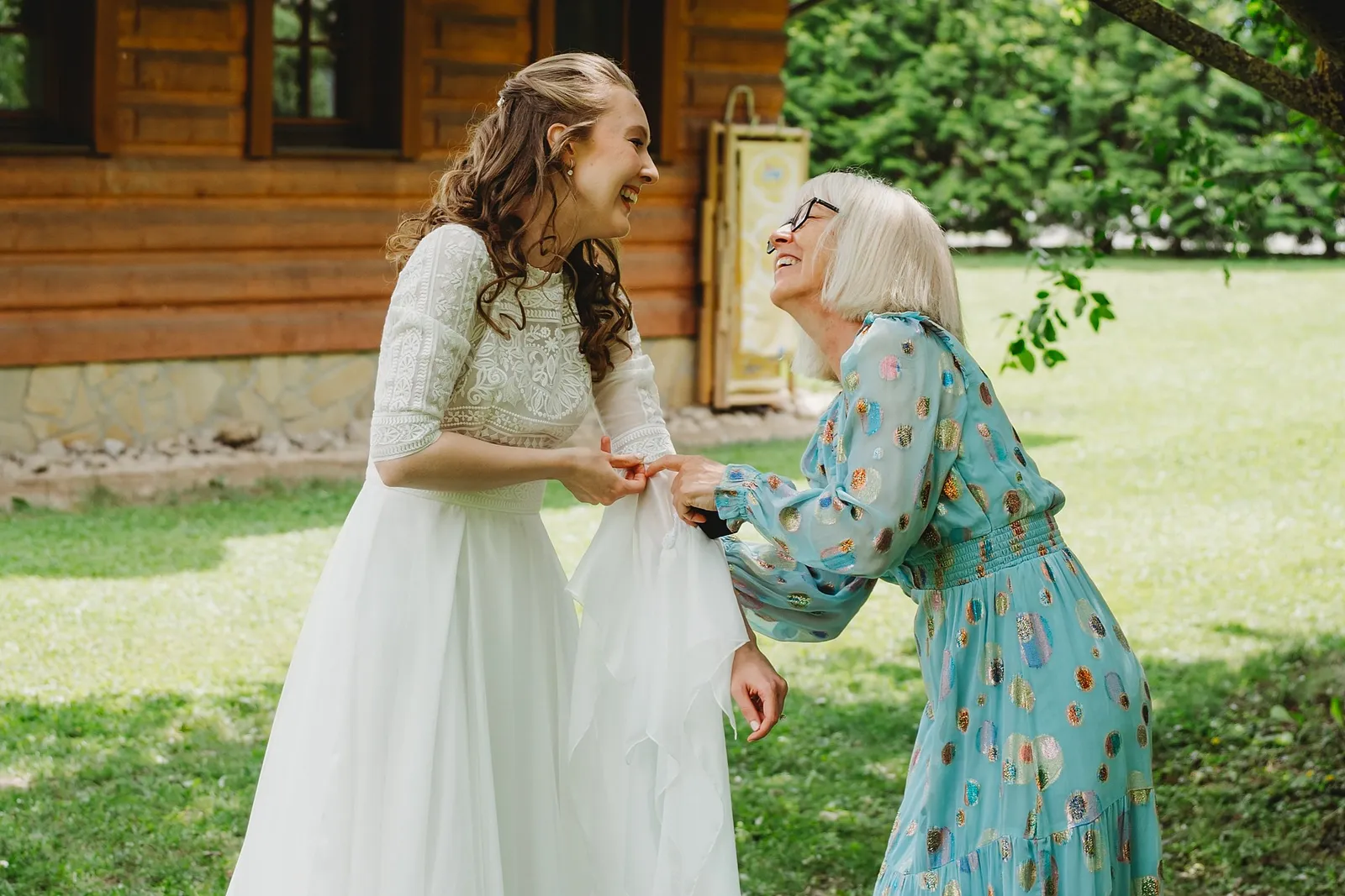 Rozosmiata žena v tyrkysových šatách pomáha neveste s rukávom šiat pred zrubom – svadba Sandra a Sebastian, Stodola Gréta