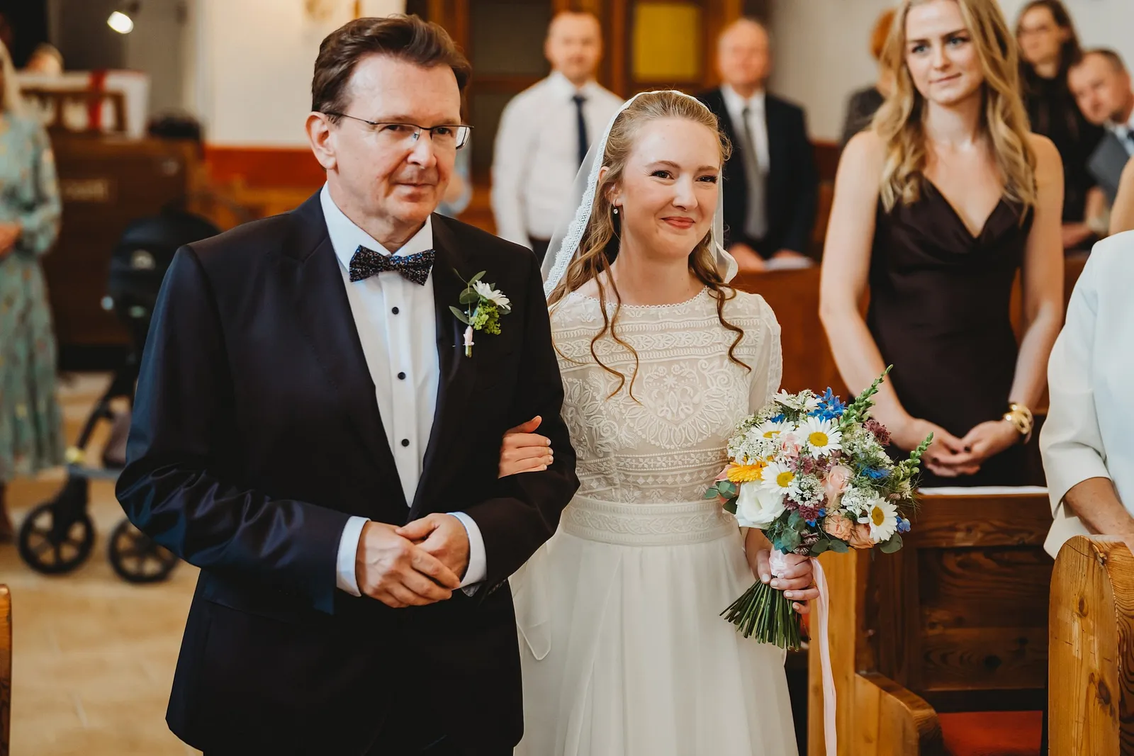 Muž v obleku s motýlikom vedie usmiatu nevestu s lúčnou kyticou uličkou kostola k oltáru – svadba Sandra a Sebastian, Stodola Gréta