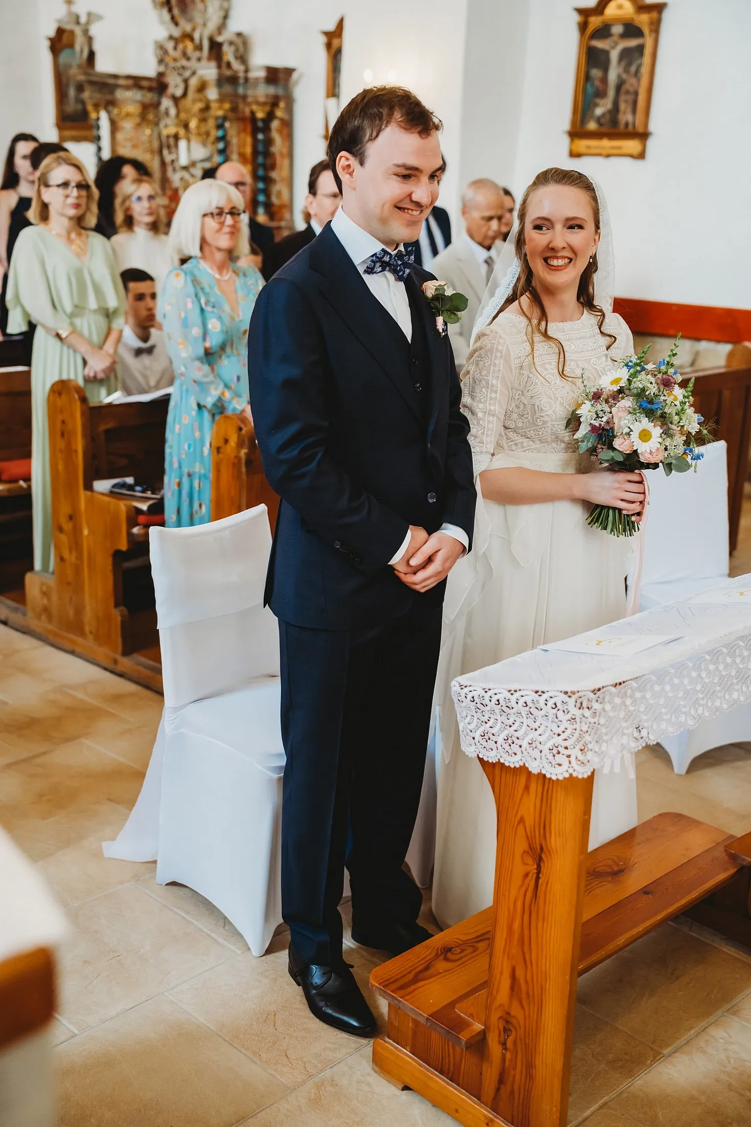 Usmiaty pár stojí pri kľakadle s čipkovou prikrývkou, nevesta drží kyticu, za nimi hostia – svadba Sandra a Sebastian, Stodola Gréta