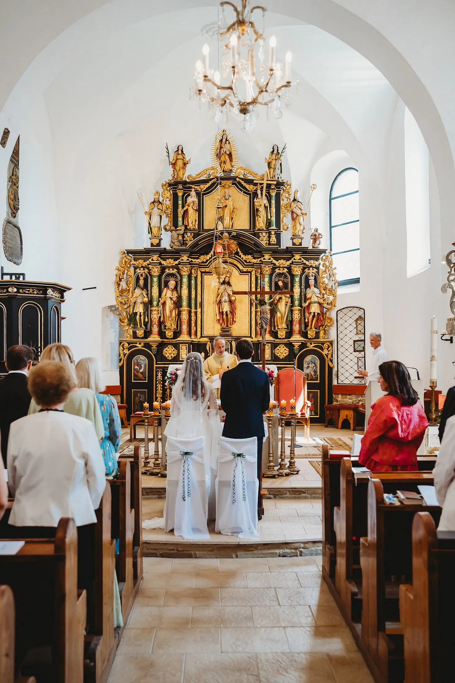 Pohľad uličkou kostola na pár pred zlatým oltárom s kňazom, nad nimi visí krištáľový luster – svadba Sandra a Sebastian, Stodola Gréta
