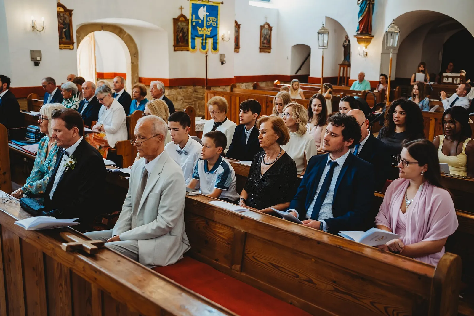 Hostia sedia v drevených laviciach kostola počas svadobného obradu – svadba Sandra a Sebastian, Stodola Gréta